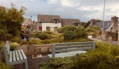 A quaint garden with green benches, surrounded by lush plants and shrubs. Stone houses with grey roofs frame the background under a cloudy sky.