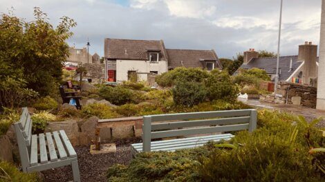 A quaint garden with green benches, surrounded by lush plants and shrubs. Stone houses with grey roofs frame the background under a cloudy sky.