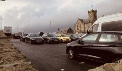 A parking lot filled with various cars, adjacent to a stone building with a clock tower and a cloudy sky above.