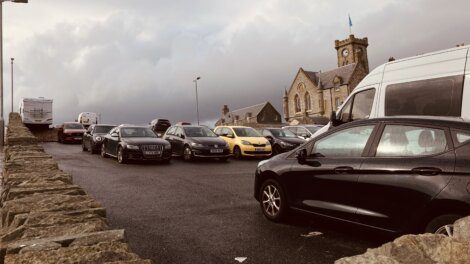 A parking lot filled with various cars, adjacent to a stone building with a clock tower and a cloudy sky above.