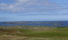 A green landscape with scattered buildings and an expanse of water in the background under a partly cloudy sky.