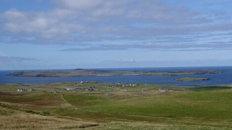 A green landscape with scattered buildings and an expanse of water in the background under a partly cloudy sky.