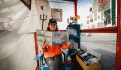 Woman in a ticket booth holding up a 'tall ship' brochure with a smile.