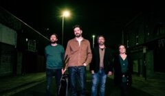 Four people stand in a dimly lit street at night. One person is holding a guitar. They all have neutral expressions and are facing the camera. The streetlights create a moody atmosphere.