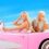 Two individuals with blond hair sit in a pink convertible car against a bright blue sky. The person driving is smiling and wearing sunglasses and colorful attire. The passenger looks at the driver.