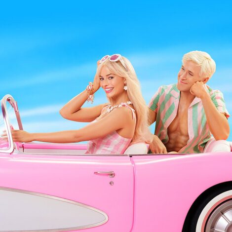 Two individuals with blond hair sit in a pink convertible car against a bright blue sky. The person driving is smiling and wearing sunglasses and colorful attire. The passenger looks at the driver.