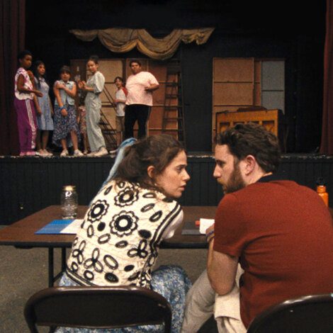 A group of people stand on stage, with one person addressing the group. Two individuals in the foreground are seated at a table, conversing and observing the stage.