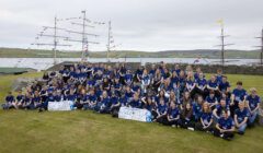 A large group of individuals wearing matching blue shirts gathers for a group photo in front of a tall ship docked at a grassy waterfront.