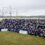 A large group of individuals wearing matching blue shirts gathers for a group photo in front of a tall ship docked at a grassy waterfront.