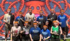 A group of people wearing orange headbands and mud-covered clothing pose in front of a "Tough Mudder Finisher" sign, holding drinks and smiling.