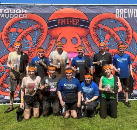 A group of people wearing orange headbands and mud-covered clothing pose in front of a "Tough Mudder Finisher" sign, holding drinks and smiling.