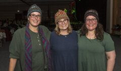 Three smiling women wearing knitted hats standing together at an event.