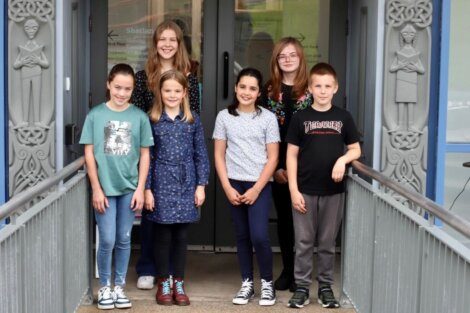 Six children stand in front of a building entrance, smiling. The entrance is flanked by decorative carvings on both sides.