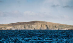 A large hill with rocky cliffs slopes gently into a blue ocean under a partly cloudy sky.