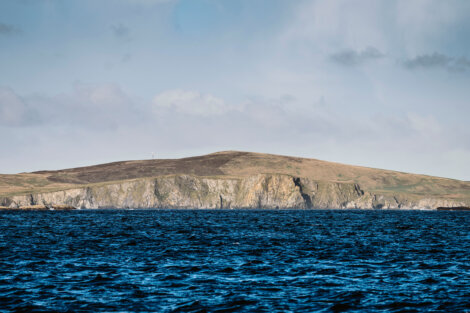 A large hill with rocky cliffs slopes gently into a blue ocean under a partly cloudy sky.