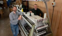 Two men smiling at the camera while standing beside an industrial embroidery machine in a workshop setting.