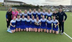 A group of female field hockey players and their coaches posing for a team photo on an astro-turf pitch, celebrating with a trophy.