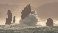 Giant waves crashing against sea stacks in a misty, stormy ocean setting.