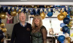 Two people smiling in a room decorated with blue and gold balloons and a banner reading "happy takeover.