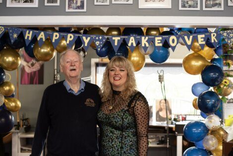 Two people smiling in a room decorated with blue and gold balloons and a banner reading "happy takeover.