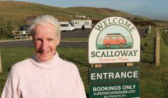 A smiling woman standing in front of a "welcome to scalloway caravan park" sign.