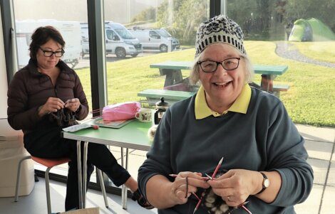 Two women knitting together at a table inside a bright room with a view of the outdoors.