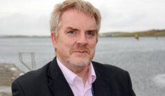 Middle-aged man with graying hair and beard standing outdoors near a body of water.