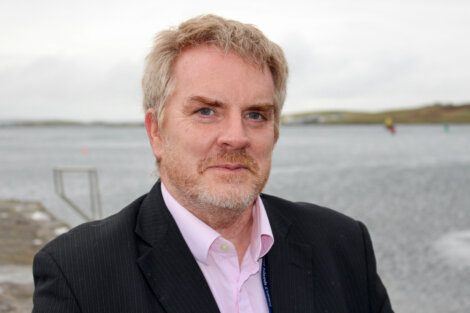 Middle-aged man with graying hair and beard standing outdoors near a body of water.