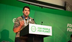 A man speaking at a podium in front of a green background.