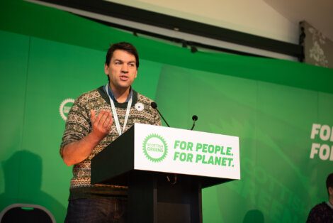 A man speaking at a podium in front of a green background.