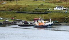 A fishing boat moored at a pier near a small white house with grassy hills in the background.