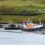 A fishing boat moored at a pier near a small white house with grassy hills in the background.