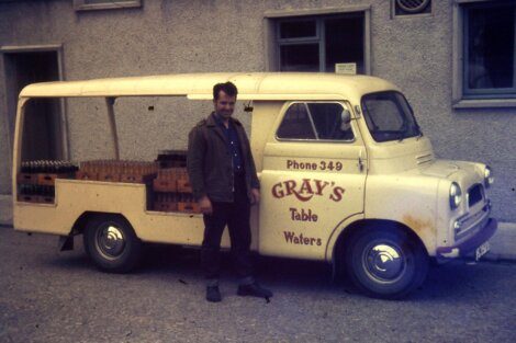 Man standing next to a vintage delivery van labeled "gray's table waters" with crates in the back.