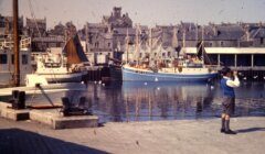 A person taking a photograph at a harbor with moored fishing boats and buildings in the background.