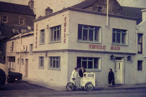 Three individuals outside the thule bar, with one on a three-wheeled cart, in a vintage street setting.