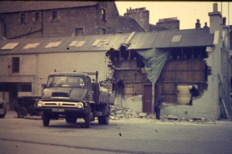 A vintage truck driving past a building with visible damage and a makeshift tarp cover.