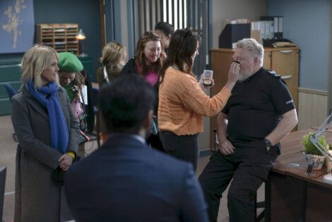 A group of people gathered around a seated man in a police uniform, with one person playfully touching his face while others look on with smiles.