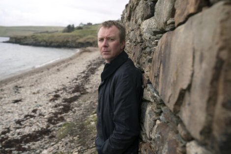 A man leaning against a stone wall near a beach under overcast skies.