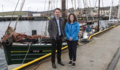 Two individuals standing by a docked sailing vessel named 'swan' at a harbor.