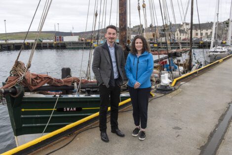 Two individuals standing by a docked sailing vessel named 'swan' at a harbor.