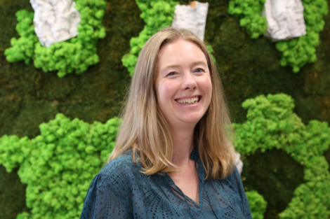 A smiling woman in a blue top standing in front of a green moss wall.