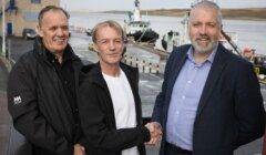 Three men shaking hands at a harbor with boats in the background.