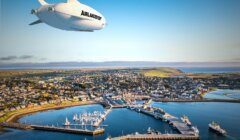 A white airship flying over a harbor and town.