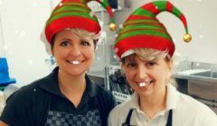 Two women wearing elf hats in a kitchen.