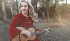 A woman is sitting in the woods with an acoustic guitar.