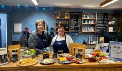 Two women standing in front of a counter with food.