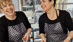 Two women smiling while preparing food in a kitchen.