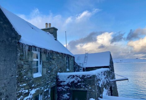 A stone house with snow on the roof next to a body of water.