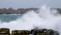 Waves crashing waves on rocks by a town with niagara falls in the background.