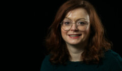 A woman with red hair and glasses is smiling in front of a black background.